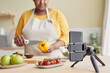 Black woman filming cooking video in kitchen, focus on smartphone at tripod, copy space