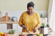 Portrait of smiling black woman cutting bananas while making healthy meal in kitchen and filming cooking video, copy space