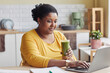 Portrait of overweight black woman drinking smoothie and using laptop at home