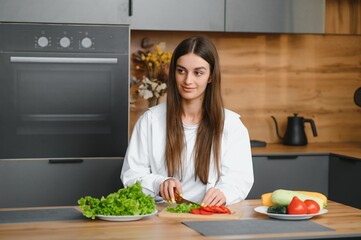 Wall Mural - Happy smiling cute woman is preparing a fresh healthy vegan salad with many vegetables in the kitchen at home and trying a new recipe