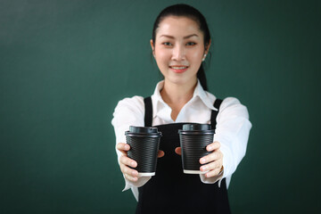 Black paper disposable coffee cups on hand of beautiful Asian female barista wearing apron, female giving two coffee cups to camera on green background, smiling pretty waitress staff working at cafe.