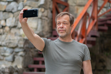 Wall Mural - Street Portrait of an elderly man 45-50 years old in a T-shirt in summer, taking a selfie on his phone against the background of an old stone wall.