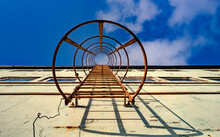 A Rusty, Vertical, Emergency Ladder Connected To A Wall Of An Abandoned Warehouse With A Blue Sky And Some Fluffy Clouds Above. 