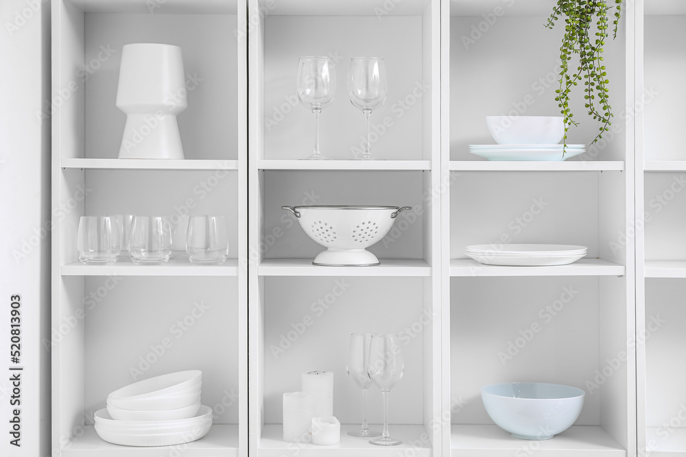 Big shelving unit with tableware near light wall
