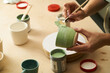 Close-up of girl painting clay mug with glaze. Woman coloring pottery in workshop with a paintbrush. Painter in green apron glazing clay pot