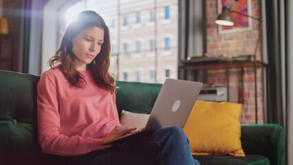 Wall Mural - Young Beautiful Female Sitting on a Couch in Loft Apartment, Working from Home on Laptop Computer. Creative Woman Checking Social Media, Browsing Internet. Urban City View from Big Window.