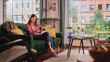 canvas print picture - Young Beautiful Female Sitting on a Couch in Living Room, Resting and Using Smartphone. Creative Girl Checking Social Media, Chatting with Friends, Browsing Internet. City View from Big Window.