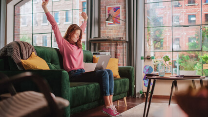 Poster - Young Beautiful Female Sitting on a Couch in Living Room, Working from Home on Laptop Computer. Creative Woman Relaxing, Stretching, Checking Social Media. Urban City View from Big Window.
