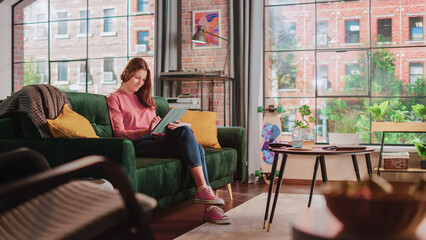 Poster - Young Beautiful Female Sitting on a Couch in Loft Apartment, Working from Home on Tablet Computer. Creative Woman Checking Social Media, Browsing Internet. Urban City View from Big Window.