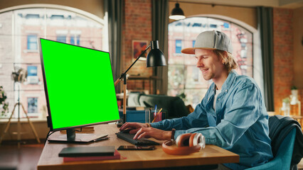 Wall Mural - Young Handsome Man Working from Home on Desktop Computer with Green Screen Mock Up Display. Creative Male Checking Social Media, Browsing Internet. Urban City View from Big Window.