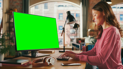 Leinwandbilder - Young Beautiful Adult Woman Working from Home on Desktop Computer with Green Screen Mock Up Display. Creative Female Checking and Writing Emails. Loft Apartment with Urban City View from Big Window.