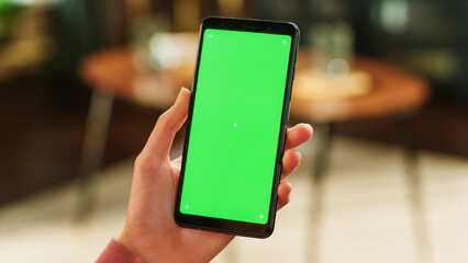 Fototapete - Feminine Hand Holding a Smartphone with Green Screen Mock Up Display. Female in Casual Clothes Resting at Home and Watching Videos on Mobile Device. Close Up Over the Shoulder Photo.