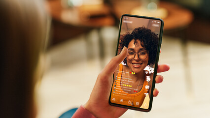 Fototapete - Feminine Hand Holding a Smartphone with Social Media Application on Display. Female in Casual Clothes Resting at Home and Watching Videos on Mobile Device. Close Up Over the Shoulder Photo.