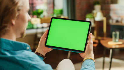 Fototapete - Young Man Holding Tablet Computer with Green Screen Mock Up Display. Male Relaxing at Home, Watching Videos and Reading Social Media Posts on Mobile Device. Close Up Over the Shoulder Photo.