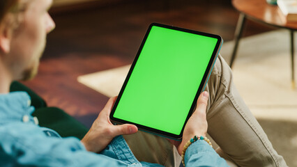 Wall Mural - Young Man Holding Tablet Computer with Green Screen Mock Up Display. Male Relaxing at Home, Watching Videos and Reading Social Media Posts on Mobile Device. Close Up Static Over the Shoulder Footage.