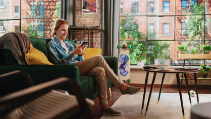 Poster - Young Handsome Adult Man Sitting on a Couch in Loft Apartment, Relaxing and Using Smartphone. Creative Male Checking Social Media, Chatting with Friends, Browsing Internet. City View from Big Window.