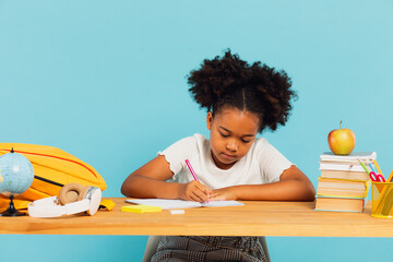 Happy African American schoolgirl doing homework at desk in class on blue background. Back to school concept.