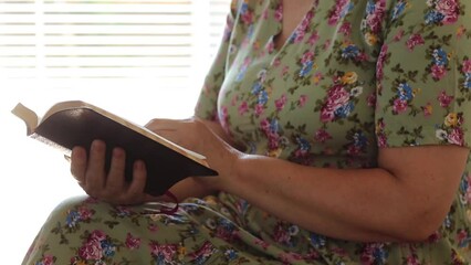 Wall Mural - A senior lady wearing a vintage dress is sitting and holding an open Holy Bible Book at home. A close-up. Faithful Christian woman read and study the Word of God Jesus Christ indoors.