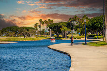 A Long Winding Footpath Along The Blue Rippling Waters Of The Lagoon With People In Swan Shaped Pedal Boats Surrounded By Lush Green Trees, Grass And Plants With Powerful Clouds At Sunset