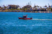 A Red And White Fire Boat Sailing On The Blue Ocean Water In The Harbor Surrounded By Lush Green Trees And Plants And Shipping Cranes In The Skyline At ShoreLine Aquatic Park In Long Beach California
