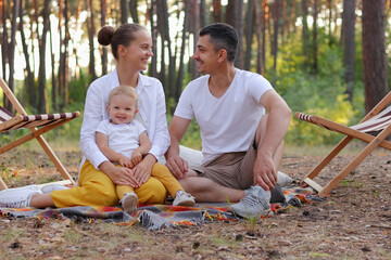 Wall Mural - Horizontal shot of happy family enjoying moment while sitting on blanket in wood with infant daughter and looking at each other with love and gentle, parents with small daughter relaxing in forest.