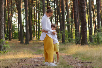 Wall Mural - Full length portrait of happy mother standing and hugging her cute charming daughter in the middle of the forest, family walking in wood, breathing fresh air, enjoying nature.