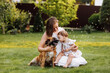 Family day, mother's day. Beautiful smiling young mom and baby daughter cuddling happy domestic dog on the backyard lawn.Mother with child girl are having fun with pet outdoors on summer holiday.