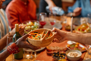 Close up of two people handing salad bowl across dinner table in cozy setting, copy space