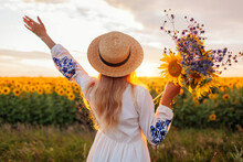 Back View Of Happy Woman Enjoying View In Blooming Sunflower Field At Sunset With Bouquet Of Flowers. Peace And Freedom