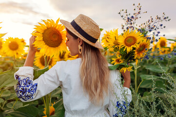 Wall Mural - Portrait of young woman walking in blooming sunflower field at sunset smelling picking flowers. Summer in Ukraine