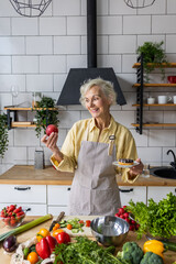 Wall Mural - Attractive smiling senior woman with apple and donut, looking happy and healthy. Concept of active lifestyle of mature people, organic food. Female at home preparing vegetarian salad, holding a cake