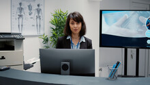 Portrait Of Receptionist Working At Hospital Reception Desk To Help People With Appointment For Checkup Visit. Waiting Area With Administrative Information To Support Healthcare System.
