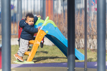 An African American Kid Dreassed In Winter Clothes Is Moving Up For A Slide In A Residence Playground Looking Into The Camera Surrounded By Metal Fence. High Quality Photo