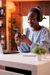 © DC Studio - Smiling african american businesswoman typing message on mobile phone. Female freelancer having break at work and watching entertaining videos on smartphone in modern home office