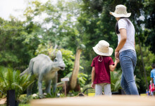 Mother To Adventure Travel In History Park With Son, Asian Child, Kid Or Little Boy. Include Dinosaur Sculpture, Grass, Field, Landscape At Outdoor Garden. Caucasian Family From Chiang Mai, Thailand.
