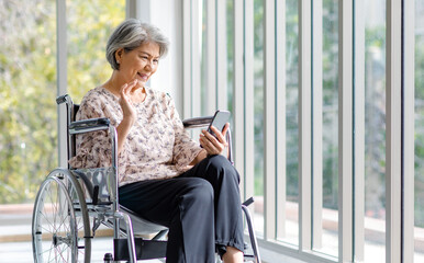 Asian happy cheerful old senior healthy gray hair retired pensioner disability handicapped grandmother sitting smiling on wheelchair using touchscreen smartphone surfing browsing social media online