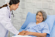 © Bangkok Click Studio - Closeup shot of unrecognizable unknown doctor in white lab coat with stethoscope hand holding comforting supporting old senior unhealthy patient in hospital uniform laying down on bed in ward room