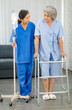 Asian young female professional doctor in uniform holding saline solution pole helping old senior gray hair pensioner patient walking with four legged assist support walker in hospital ward room