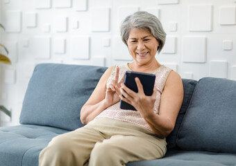 Asian cheerful old senior healthy gray hair female pensioner grandmother sitting smiling on cozy sofa in living room holding using touchscreen tablet computer surfing browsing application online