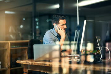 Hes not afraid of hard work. Shot of a young businessman talking on his phone and using a computer during a late night at work.