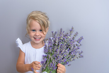 Joyful laughing white caucasian girl 2 years old with blond hair with a bouquet of lavender in her hands in a white dress on a gray background during the holiday