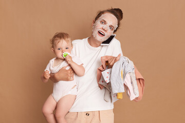 Wall Mural - Amazed woman wearing white t shirt doing cosmetic procedures, holding baby daughter in hands and kid's clothing, talking via cell phone with her friend, isolated over brown background.