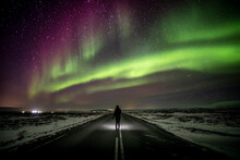 Traveler On Asphalt Road Admiring Aurora Borealis In Night Sky