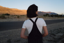 Back View Of Slim Person In Vintage Overalls With Short Hair Looking At Dusty Road In Rural Landscape