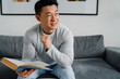 © Drobot Dean - Brunette adult asian man reading book while sitting on sofa at home