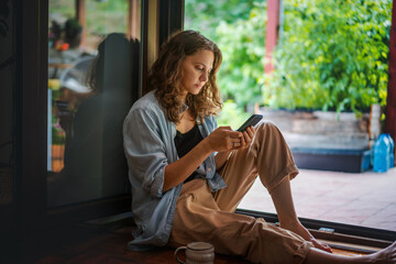 Young beautiful woman with a smartphone sitting on the wooden floor near open door to the terrace of a country house