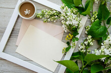 Close-up Flatlay With A Cup Of Coffee In White Wooden Frame And Mockup And Copyspace Pastel Pink Envelopes And Blank Postcard Framed By Branches Of Burgundy And Green Leaves, White Lilac Flowers 