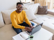 © Jelena Stanojkovic - Smiling black man using laptop at home in living room. Happy young businessman send email and working at home. African American freelancer typing on computer with notebook on table.