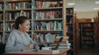 © silverkblack - Young beautiful smiling caucasian female student is sitting at desk in big spacious library with pile of books on edge. She is writing something in copybook, laptop is in front of her