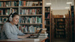 © silverkblack - Beautiful positive caucasian female student with big white headphones on head working at table in spacious library in front of laptop. She is smiling and texting